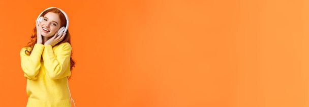 Taille-up portret schattige tiener roodharige meisje in witte hoofdtelefoon, kantelhoofd luisteren muziek, aanraking oortelefoons en glimlachende camera, meezingen favoriete lied, staande oranje achtergrond. Kopieerruimte - Foto, afbeelding