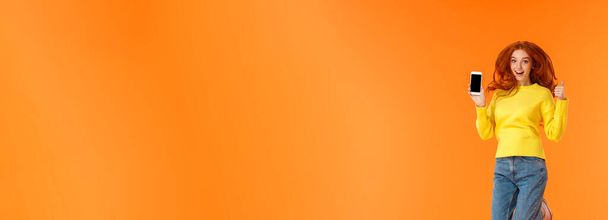 Publicité, technologie et concept de vacances. Vertical pleine longueur plan gingembre gai magnifique fille, rousse femme sautant sur fond orange avec smartphone, montrer pouce vers le haut
. - Photo, image