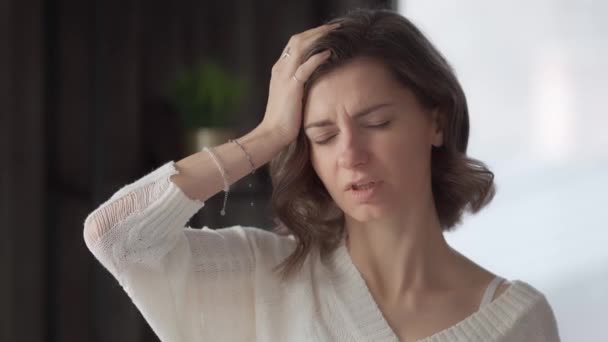 Egy fiatal nő otthon megérinti a homlokát a kezével, hogy ellenőrizze a testhőmérsékletét, fejfájástól szenvedve. Migrénroham egy felnőttnél. A légzőszervi vírusbetegség első tünetei. - Felvétel, videó