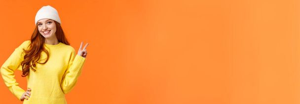 modernes Hipstermädchen, das während der Winterferien mit Freunden Schneebälle spielt, ein Friedenszeichen als Posing-Foto in der Nähe eines Schneemanns zeigt, weiße Mütze und Pullover über orangefarbenem Hintergrund trägt. - Foto, Bild