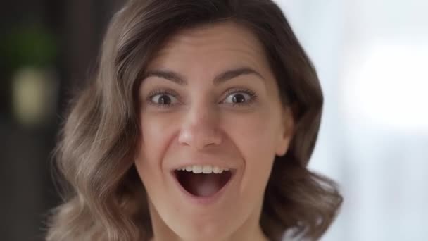 A young woman at home looks emotionally and enthusiastically into the camera, smiles happily, rejoices and laughs. Portrait of a person. Female face close-up. Small depth of field blurred background - Footage, Video