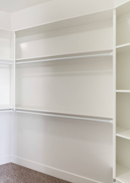 Vertical White built in wall mount shelving unit in a walk in closet. Walk in closet interior with brown carpeted flooring and framed shelves with plastic rods below. - Photo, Image