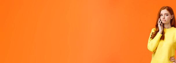Meisje dat angstaanvallen krijgt. Verbijsterd of nerveus schattig roodharige vrouw bijten lip en weg te kijken als het hebben van gesprek op mobiele telefoon, staande oranje achtergrond verdrietig. - Foto, afbeelding