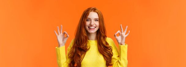 ファッション、ショッピング、冬の休日の概念。良いジェスチャーや笑顔を示す魅力的な陽気な赤毛の女性,すべてが大丈夫,承認または製品のように,オンラインストアをお勧めします,オレンジの背景. - 写真・画像