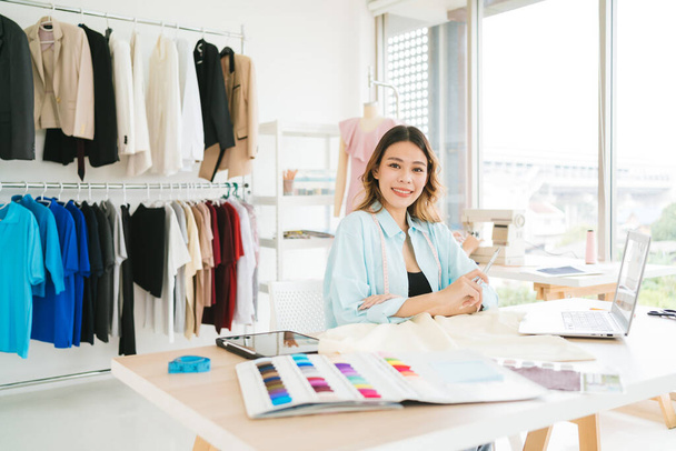 Portret pewnej siebie azjatyckiej krawcowej kobiety pracującej nad ubraniami w pracowni krawieckiej. Młoda, atrakcyjna projektantka mody uśmiecha się i patrzy w kamerę z manekinem w tle. - Zdjęcie, obraz