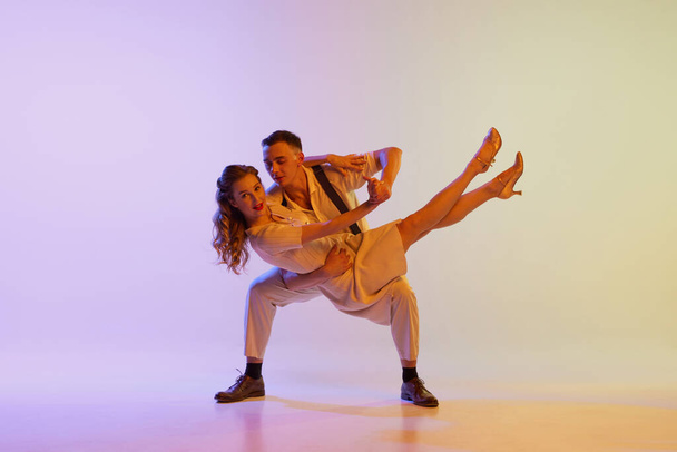 Beautiful girl and man in retro style costumes dancing incendiary dances isolated on gradient lilac color background in neon light. Dancers in motion and action. Concept of art, 60s, 70s culture - Photo, Image