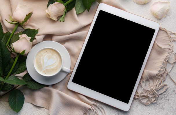 Spazio di lavoro minimalista con tablet digitale mockup schermo vuoto, sciarpa accogliente, una tazza di caffè e fiori di rose su sfondo di cemento. Vista dall'alto, piatta - Foto, immagini