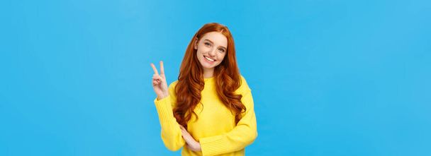 Waist-up shot cheerful tender and cute redhead modern woman in yellow sweater, tilt head and smiling lovely, showing peace sign, express positive emotions, joy and happiness, blue background. - Photo, Image