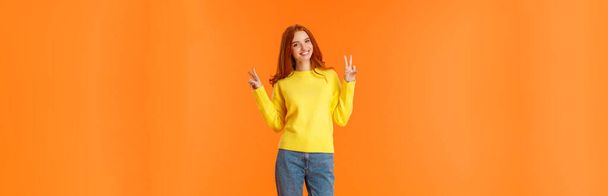 Vertical full-length bonito linda ruiva adolescente em jeans e camisola amarela de inverno de pé com sinais de paz sobre fundo laranja, posando sorridente expressar emoções alegrias despreocupadas
. - Foto, Imagem