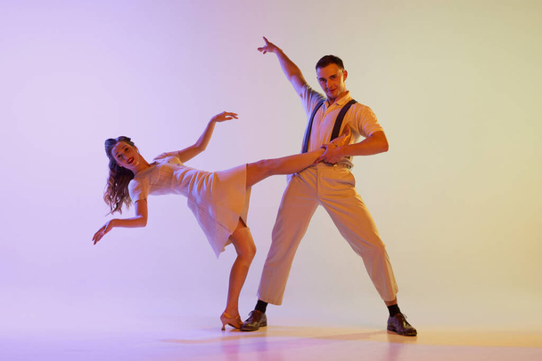 Musical show. Emotional couple of dancers in retro style outfits dancing social dances isolated on gradient lilac color background in neon light. Dancers in motion and action. - Φωτογραφία, εικόνα