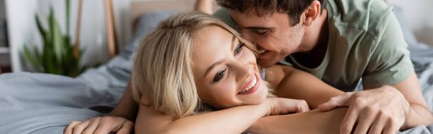 Uśmiechnięty młody mężczyzna całujący blondynkę na łóżku rano, baner  - Zdjęcie, obraz
