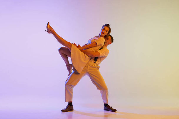 Beautiful girl and man in retro style costumes dancing incendiary dances isolated on gradient lilac color background in neon light. Dancers in motion and action. Concept of art, 60s, 70s culture - Fotografie, Obrázek
