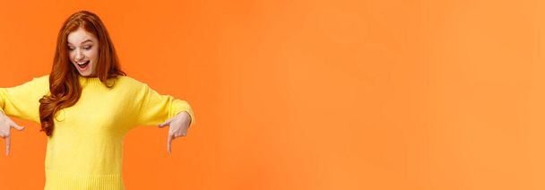 Merveilleux et excité, amusé femme rousse mignonne en pull jaune pointant les doigts vers le bas, regardez en bas et souriant, check-out promo produit, événement de vacances d'hiver, fond orange
. - Photo, image
