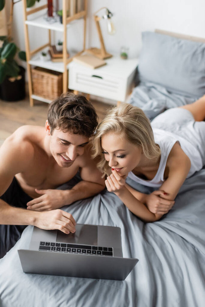 High angle view of smiling shirtless man using laptop near girlfriend in pajama on bed  - Foto, Bild