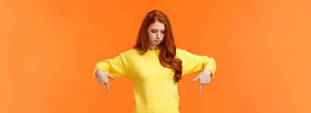 Envy, sad and sulky cute redhead woman in yellow sweater, looking and pointing down with jealousy or disappointment, feeling jealousy and regret missing chance, standing orange background. - Foto, Imagen