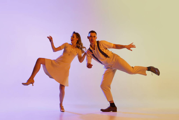 Beautiful girl and man in retro style costumes dancing incendiary dances isolated on gradient lilac color background in neon light. Dancers in motion and action. Concept of art, 60s, 70s culture - Valokuva, kuva