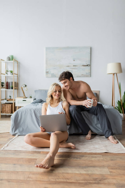 glimlachende shirtloze man met kopje koffie in de buurt van vriendin met laptop in de slaapkamer  - Foto, afbeelding