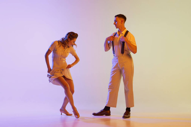 Beautiful girl and man in retro style costumes dancing incendiary dances isolated on gradient lilac color background in neon light. Dancers in motion and action. Concept of art, 60s, 70s culture - Фото, зображення