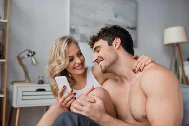 Usmívající se blondýna žena drží smartphone a objímání bez trička přítel v ložnici  - Fotografie, Obrázek