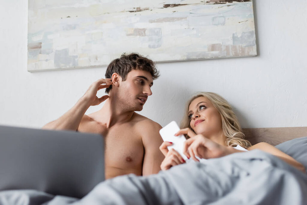 Σέξι άντρας μιλάει με τη φίλη του με το smartphone κοντά θολή φορητό υπολογιστή στο κρεβάτι  - Φωτογραφία, εικόνα