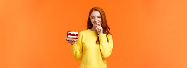 陽気で満足し、幸せな赤毛の女性がカンニングしている日おいしい食べ物を食べて、おいしい作品ケーキを保持し、唇をかむと笑顔、誘惑に抵抗することはできません、欲望、オレンジの背景をかむ. - 写真・画像