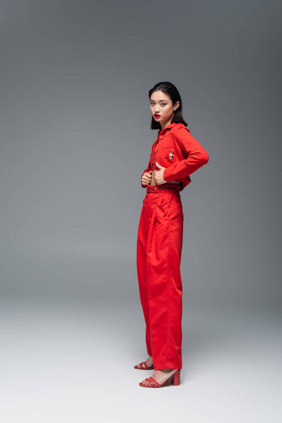full length of young asian woman in red jacket and trousers looking at camera on grey background - Фото, зображення