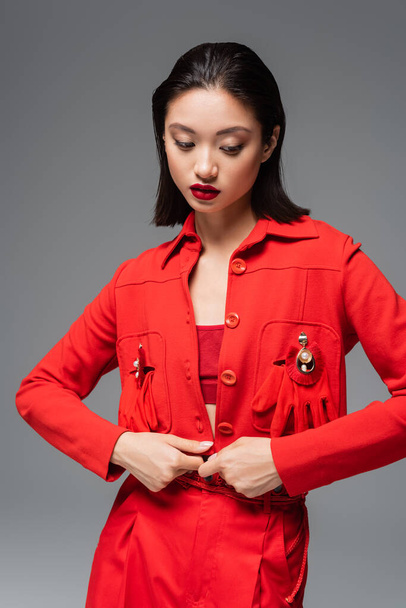 young asian woman in red jacket decorated with brooches and gloves isolated on grey - Photo, Image