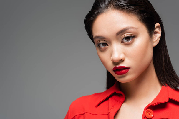 portrait of brunette asian woman with makeup and red lips looking at camera isolated on grey - Foto, afbeelding