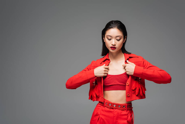 brunette asian woman touching red fashionable jacket isolated on grey - Фото, изображение