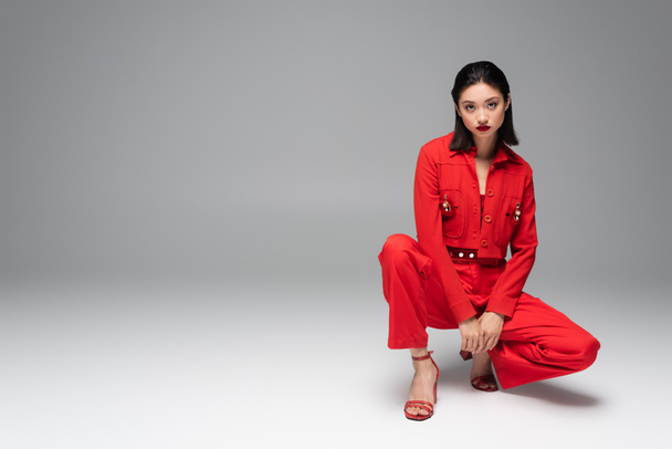 nuori aasialainen malli punainen tyylikäs puku ja sandaalit poseeraa haunches harmaalla taustalla - Valokuva, kuva