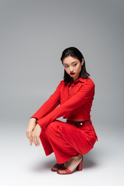 pretty asian woman in red stylish outfit sitting on haunches on grey background - Valokuva, kuva