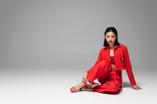 morena mujer asiática en pantalones rojos y chaqueta sentado sobre fondo gris - Foto, imagen