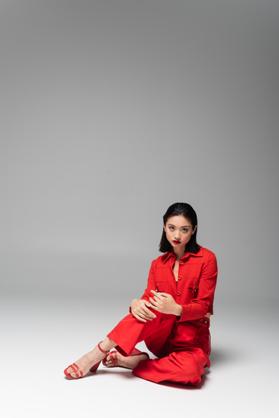brünettes asiatisches Model in roter Jacke und Hose schaut in die Kamera, während es auf grauem Hintergrund sitzt - Foto, Bild