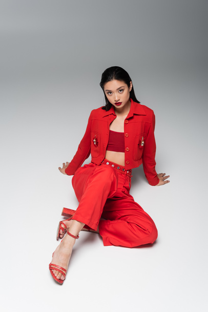 sensual asian woman in red suit and heeled sandals sitting on grey background - Fotoğraf, Görsel