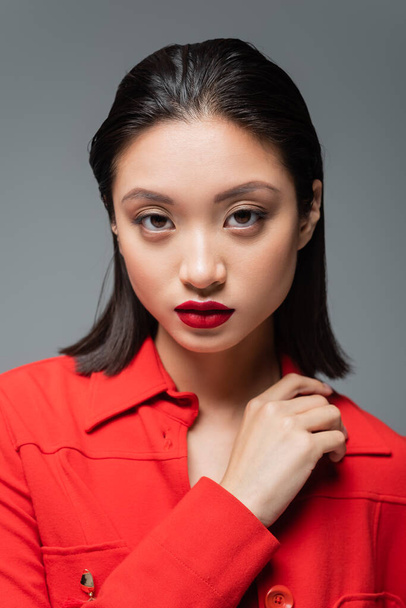 portrait of brunette asian woman with red lips wearing red blazer and looking at camera isolated on grey - Photo, Image