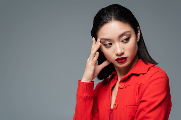 portrait of brunette asian woman in red jacket holding hand near face and looking away isolated on grey - Fotoğraf, Görsel