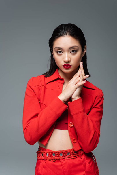 brunette asian model in red fashionable jacket looking at camera isolated on grey - Photo, Image