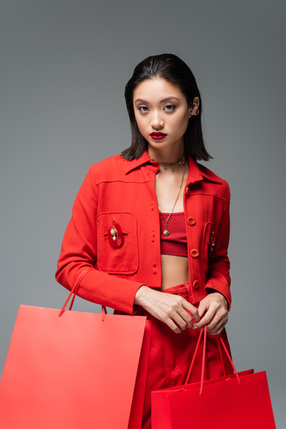 donna asiatica alla moda in giacca rossa con borse della spesa e guardando la fotocamera isolata sul grigio - Foto, immagini