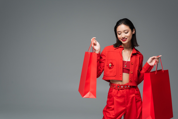 happy asian woman in elegant clothes posing with red shopping bags isolated on grey - Photo, Image