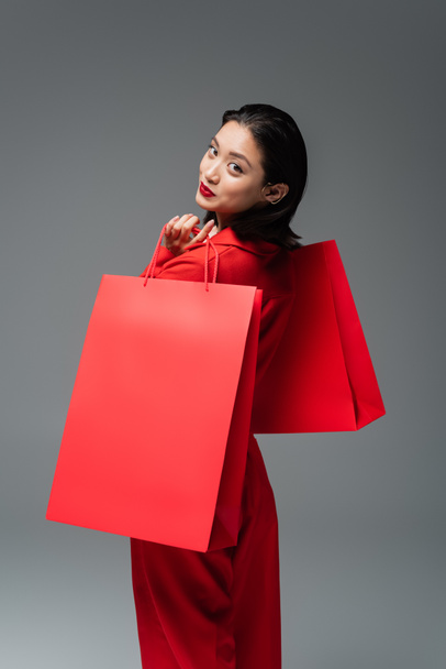 brunette asian woman with red shopping bags smiling at camera isolated on grey - Foto, immagini