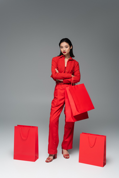 täyspitkä aasialainen nainen punainen tyylikäs puku seisoo ristissä kädet lähellä ostoskassit harmaalla pohjalla - Valokuva, kuva