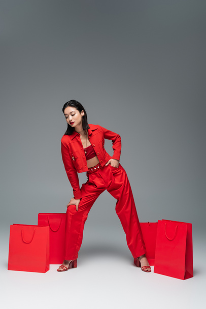 full length of asian woman in red stylish suit posing with hand in pocket near shopping bags on grey background - Fotografie, Obrázek