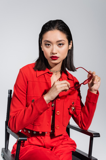 young asian woman with red lips holding stylish sunglasses while sitting isolated on grey - Foto, immagini