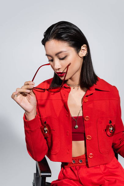 brunette asian woman in red jacket holding fashionable sunglasses isolated on grey - Foto, Imagem