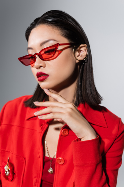 portrait of asian woman in red stylish sunglasses and ear cuff touching chin isolated on grey - Zdjęcie, obraz