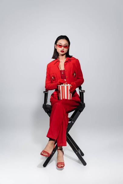 full length of asian woman in trendy sunglasses and red elegant suit sitting with popcorn bucket on grey background - Foto, immagini
