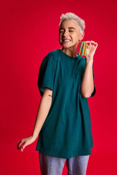 Junge stilvolle Frau mit kurzen Haaren posiert in lässiger Kleidung und isst saure Gelee Bonbons isoliert vor rotem Hintergrund. Konzept der Jugend, Schönheit, Mode, Lifestyle, Emotionen, Mimik. Anzeige - Foto, Bild