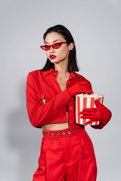 asian woman in trendy sunglasses and red outfit holding bucket of popcorn and looking away on grey background - Фото, изображение