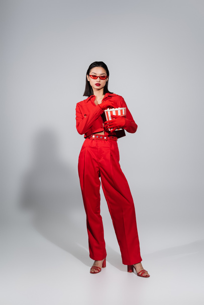 full length of asian woman wearing sunglasses and red suit with gloves holding popcorn bucket on grey background - Photo, Image