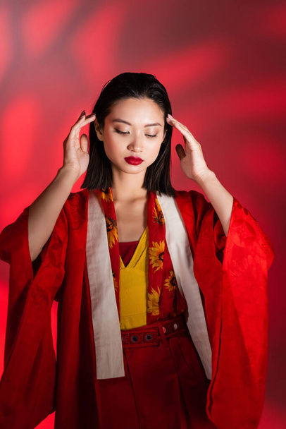 elegant asian woman in red kimono cape posing with hands near head on abstract background - Fotó, kép